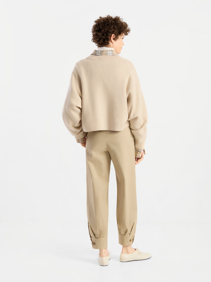 Eine Person mit kurzen, lockigen Haaren, die den Pullover „Giulia Puder“ aus Merinowolle über einem karierten Hemd trägt, dazu eine hellbraune Hose mit geknöpften Bündchen und beige Schuhe, steht mit dem Rücken zur Kamera in einem weißen Studio.