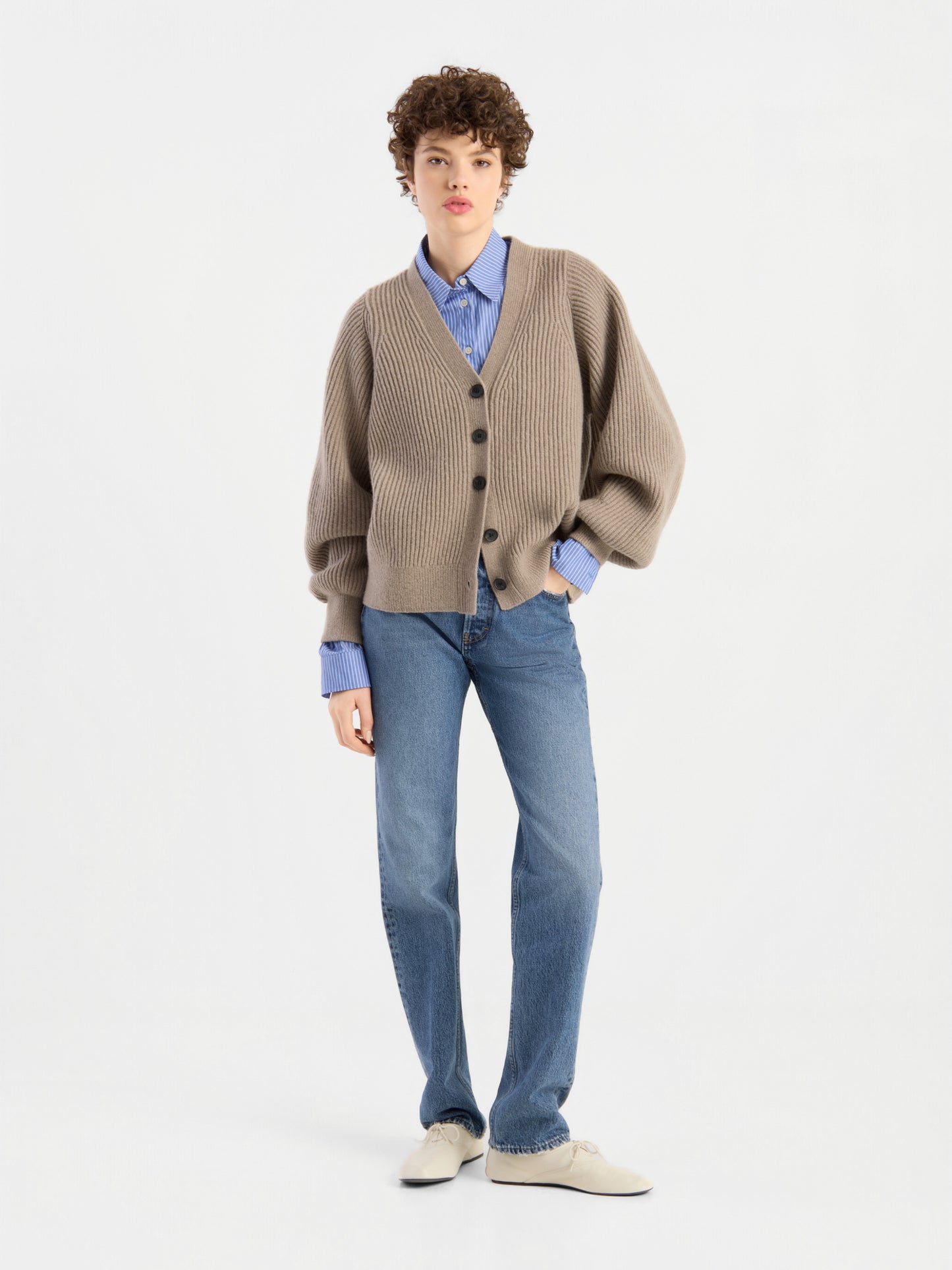 Eine Person mit kurzen, lockigen Haaren trägt die Strickjacke „Antonia Rib Beige“ mit einer weichen Rippenstruktur über einem blauen Hemd, blauen Jeans und weißen Schuhen und steht vor einem schlichten weißen Hintergrund.