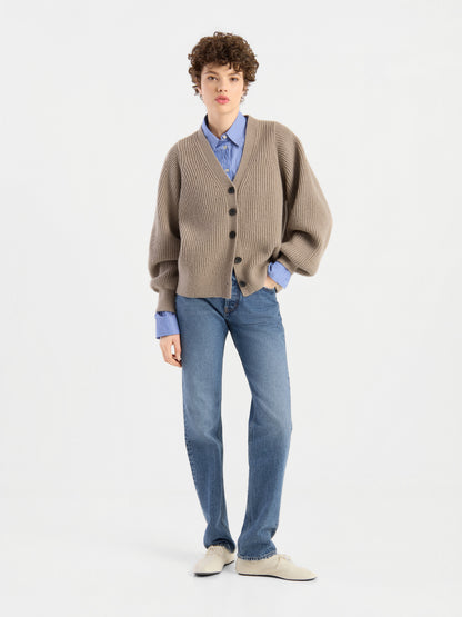Eine Person mit kurzen, lockigen Haaren trägt die Strickjacke „Antonia Rib Beige“ mit einer weichen Rippenstruktur über einem blauen Hemd, blauen Jeans und weißen Schuhen und steht vor einem schlichten weißen Hintergrund.