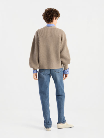 Eine Person mit lockigem Haar steht mit dem Rücken zur Kamera und trägt die Strickjacke „Antonia Rib Beige“ mit sanfter Rippenstruktur, über einem hellblauen Hemd mit Kragen, dazu blaue Jeans und weiße Schuhe vor einem schlichten weißen Hintergrund.