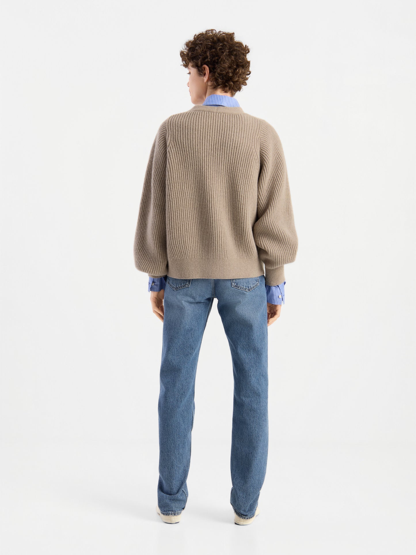 Eine Person mit kurzen Locken ist von hinten zu sehen. Sie trägt die Strickjacke „Antonia Rib Beige“ – eine taupefarbene Strickjacke mit dezenter Rippenstruktur –, die über einem blauen Hemd, einer blauen Jeans und weißen Schuhen vor einem schlichten weißen Hintergrund liegt.