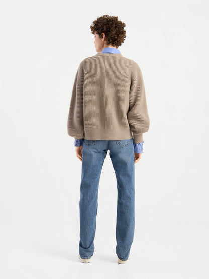Eine Person mit kurzen Locken ist von hinten zu sehen. Sie trägt die Strickjacke „Antonia Rib Beige“ – eine taupefarbene Strickjacke mit dezenter Rippenstruktur –, die über einem blauen Hemd, einer blauen Jeans und weißen Schuhen vor einem schlichten weißen Hintergrund liegt.