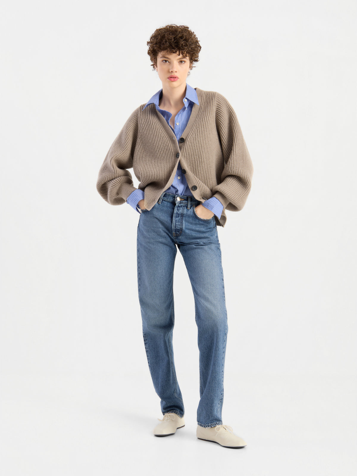 Eine Person mit kurzen Locken präsentiert die Strickjacke „Antonia Rib Beige“ mit ihrer eleganten Rippenstruktur, kombiniert mit einem blauen Hemd, blauen Jeans und weißen Schuhen vor einem schlichten weißen Hintergrund.
