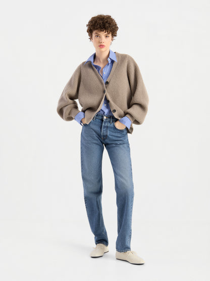 Eine Person mit kurzen Locken präsentiert die Strickjacke „Antonia Rib Beige“ mit ihrer eleganten Rippenstruktur, kombiniert mit einem blauen Hemd, blauen Jeans und weißen Schuhen vor einem schlichten weißen Hintergrund.
