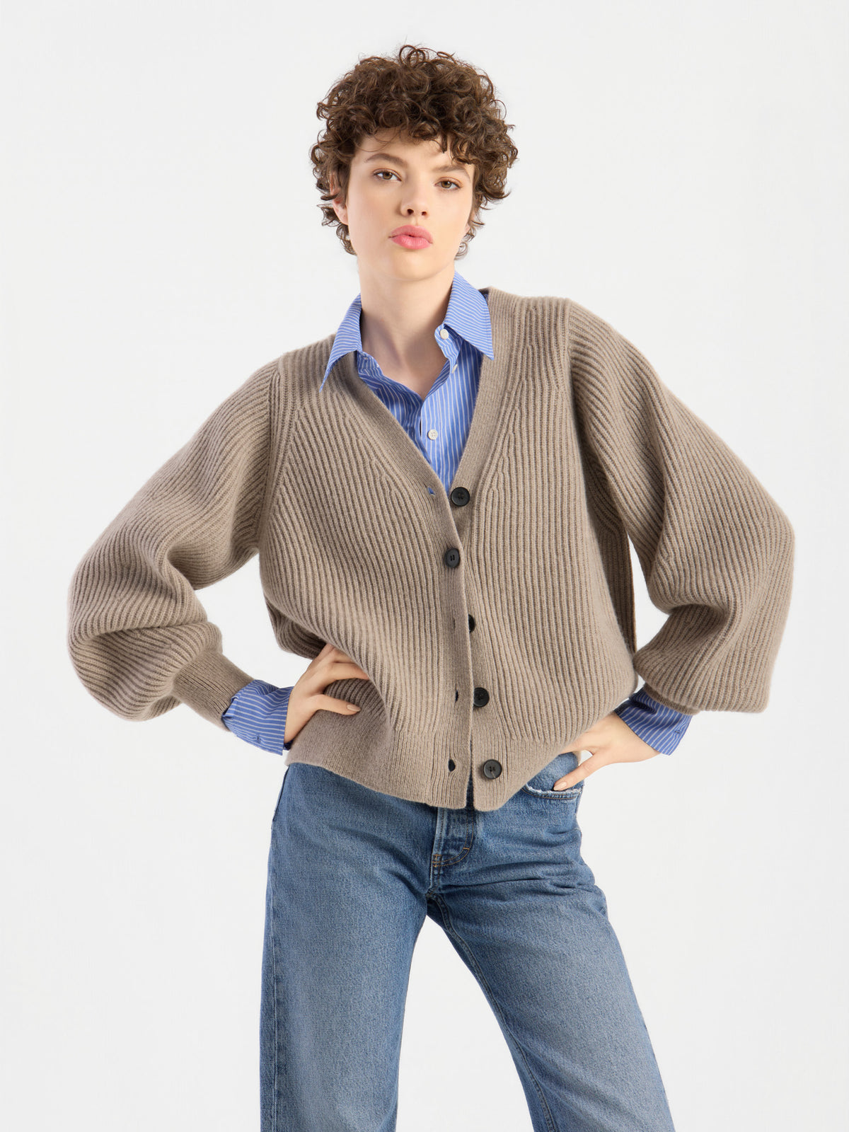 Eine Person mit kurzen Locken präsentiert die Strickjacke „Antonia Rib Beige“ mit ihrer markanten Rippenstruktur. Sie trägt dazu ein blaues Hemd mit Kragen und eine blaue Jeans und steht mit einer Hand in der Hüfte vor einem schlichten weißen Hintergrund.