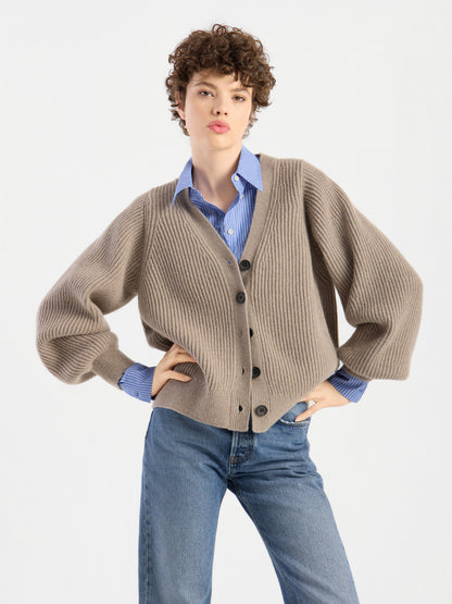 Eine Person mit kurzen Locken präsentiert die Strickjacke „Antonia Rib Beige“ mit ihrer markanten Rippenstruktur. Sie trägt dazu ein blaues Hemd mit Kragen und eine blaue Jeans und steht mit einer Hand in der Hüfte vor einem schlichten weißen Hintergrund.