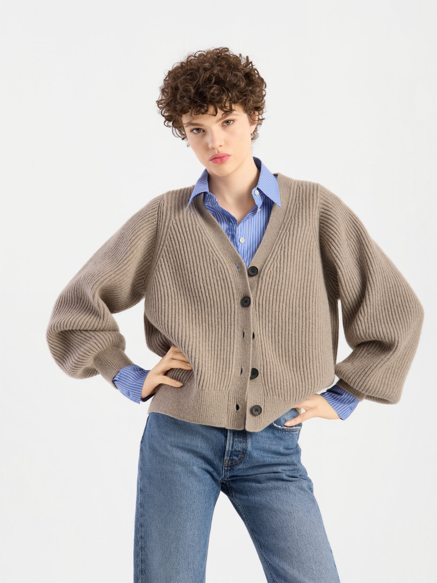 Eine Person, die die Strickjacke „Antonia Rib Beige“ – eine beige gerippte Damenstrickjacke – über einem blau gestreiften Hemd und einer blauen Jeans trägt, steht mit einer Hand in der Hüfte vor einem schlichten, hellen Hintergrund.