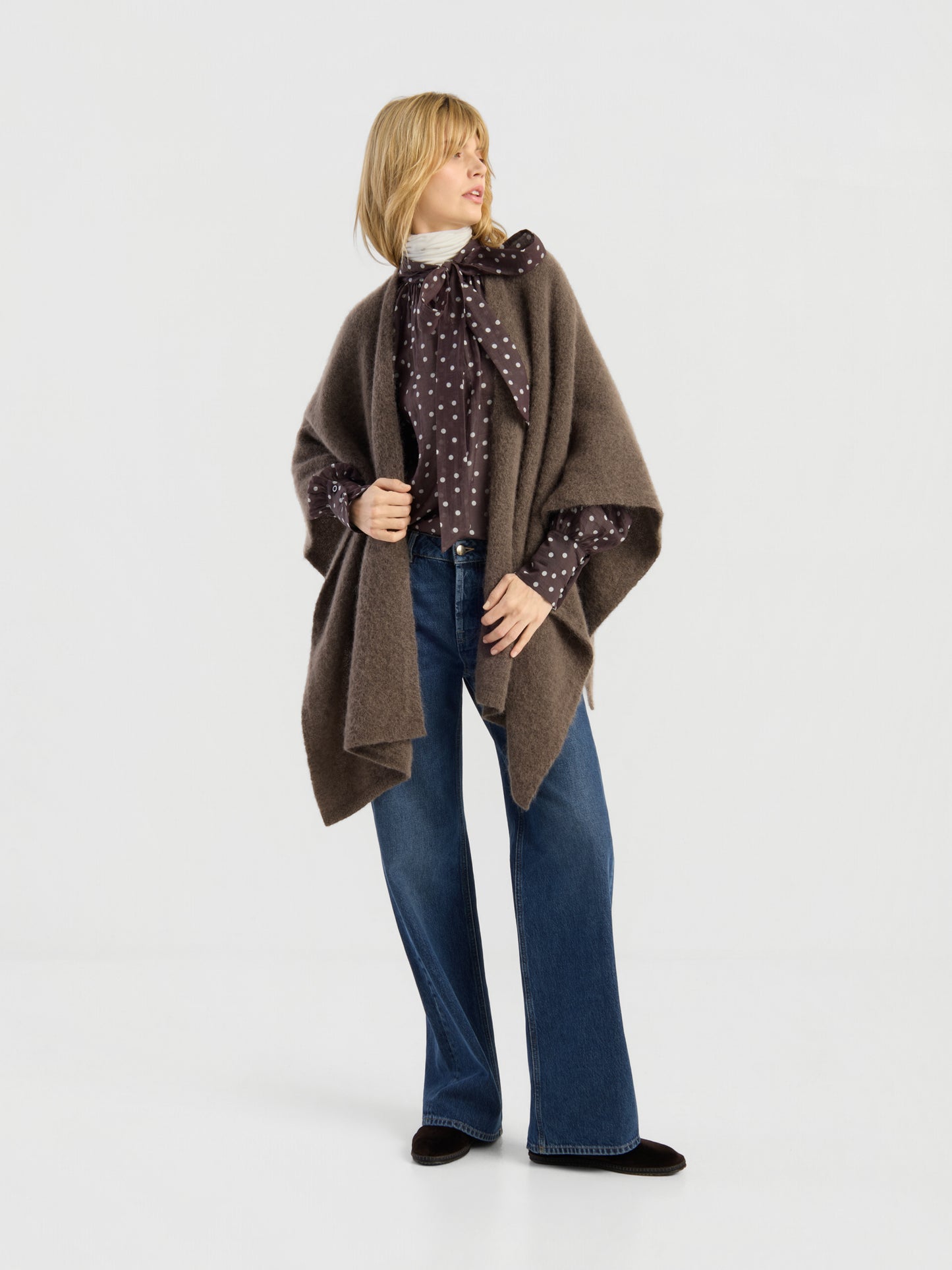 Eine Person, die das „Bear Cape Braun“, eine gepunktete Bluse mit Schleife, weit geschnittene Jeans und schwarze Schuhe trägt, steht in einer entspannten Haltung vor einem schlichten, hellen Hintergrund.