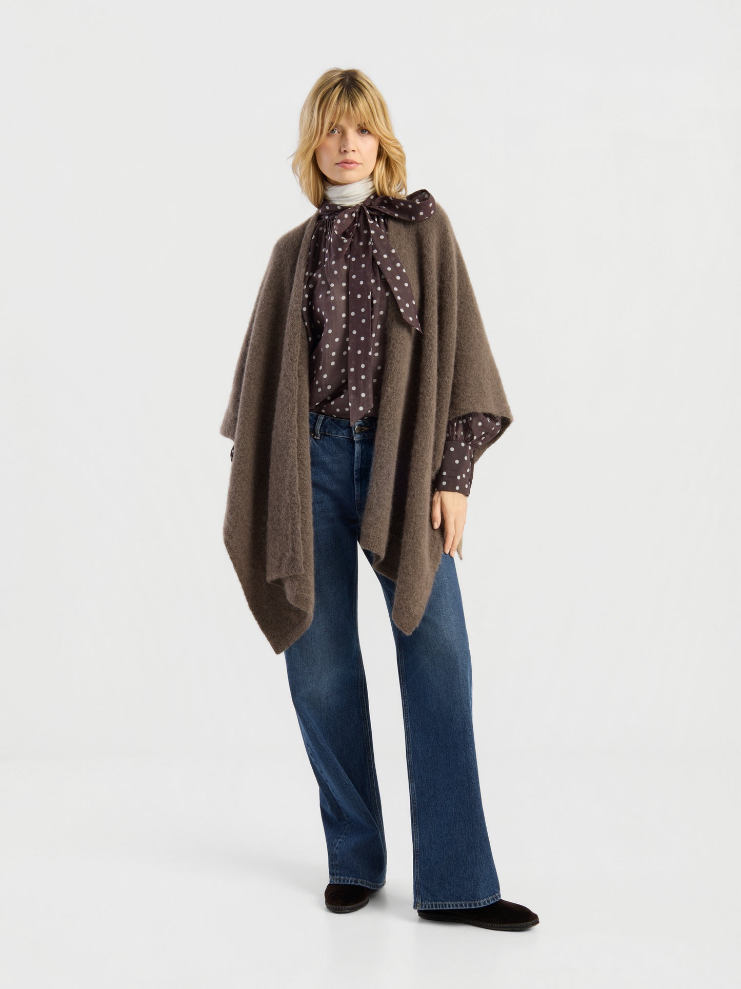 Vor einem schlichten weißen Hintergrund steht eine Frau, die das „Bear Cape Braun“ über einer gepunkteten Bluse mit Schleife, einer blauen Jeans mit weitem Bein und schwarzen Schuhen trägt.