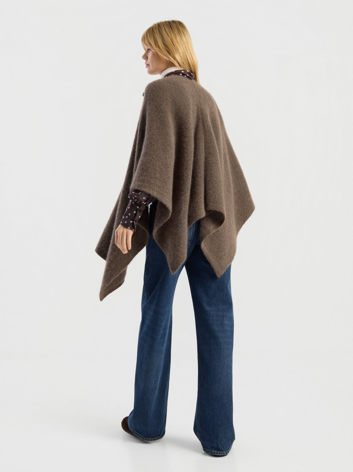 Eine Frau mit glattem blondem Haar steht mit dem Rücken zur Kamera vor einem schlichten weißen Hintergrund und trägt das „Bear Cape Braun“ über einem dunkel gemusterten Shirt und einer blau gemusterten Jeans mit weitem Bein.