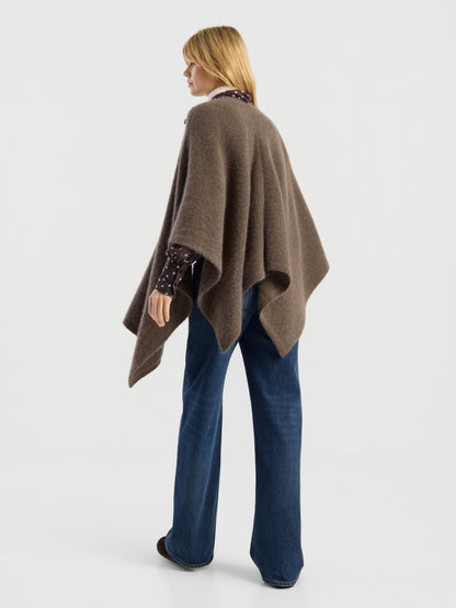 Eine Frau mit glattem blondem Haar steht mit dem Rücken zur Kamera vor einem schlichten weißen Hintergrund und trägt das „Bear Cape Braun“ über einem dunkel gemusterten Shirt und einer blau gemusterten Jeans mit weitem Bein.