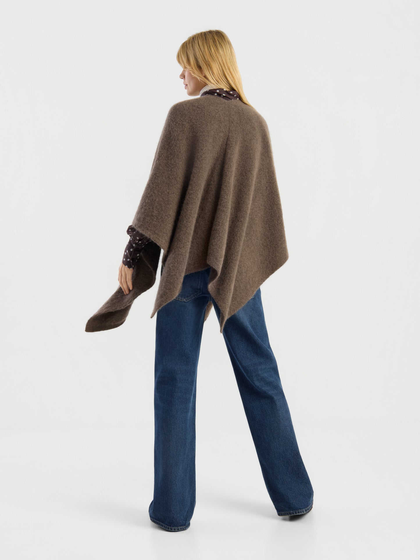 Eine Person mit glattem blondem Haar, die den „Bear Cape Braun“ über einem gemusterten Hemd und blauen Jeans trägt, steht vor einem schlichten weißen Hintergrund und wendet der Kamera den Rücken zu.