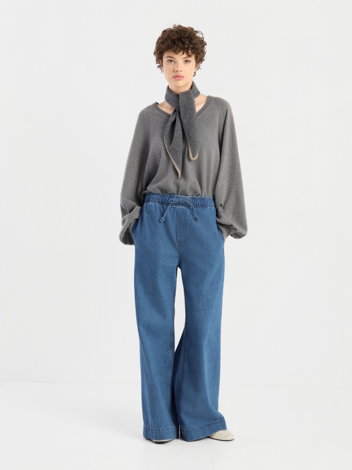 Eine Person trägt einen locker sitzenden grauen Pullover mit Schal-Detail und die Damenjeans „Anna Pants“ mit elastischem Bund vor einem schlichten weißen Hintergrund.
