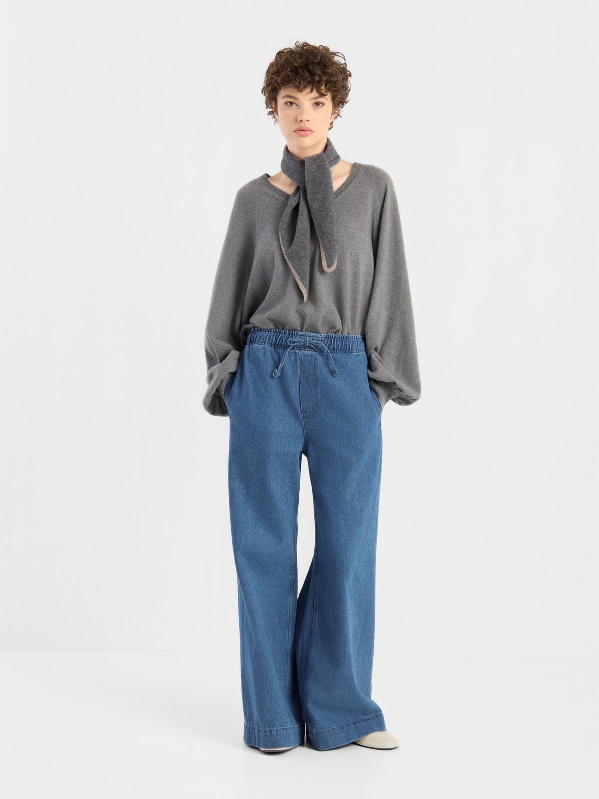 Eine Person, die einen grauen Pullover trägt, an dessen Hals der Schal „Sophie Grau“ als Halstuch gebunden ist, sowie eine locker sitzende blaue Jeans, steht vor einem schlichten weißen Hintergrund.