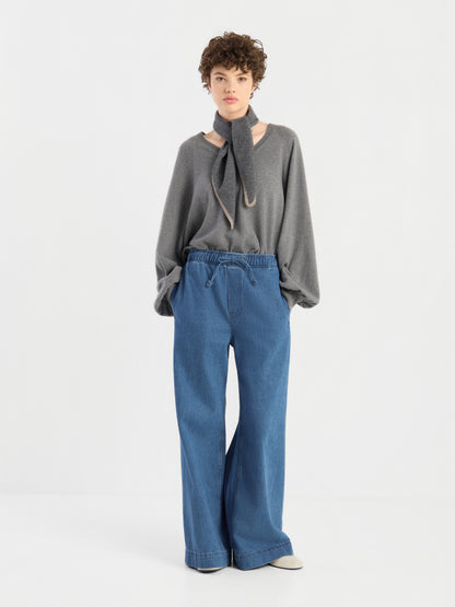Eine Person, die einen grauen Pullover trägt, an dessen Hals der Schal „Sophie Grau“ als Halstuch gebunden ist, sowie eine locker sitzende blaue Jeans, steht vor einem schlichten weißen Hintergrund.