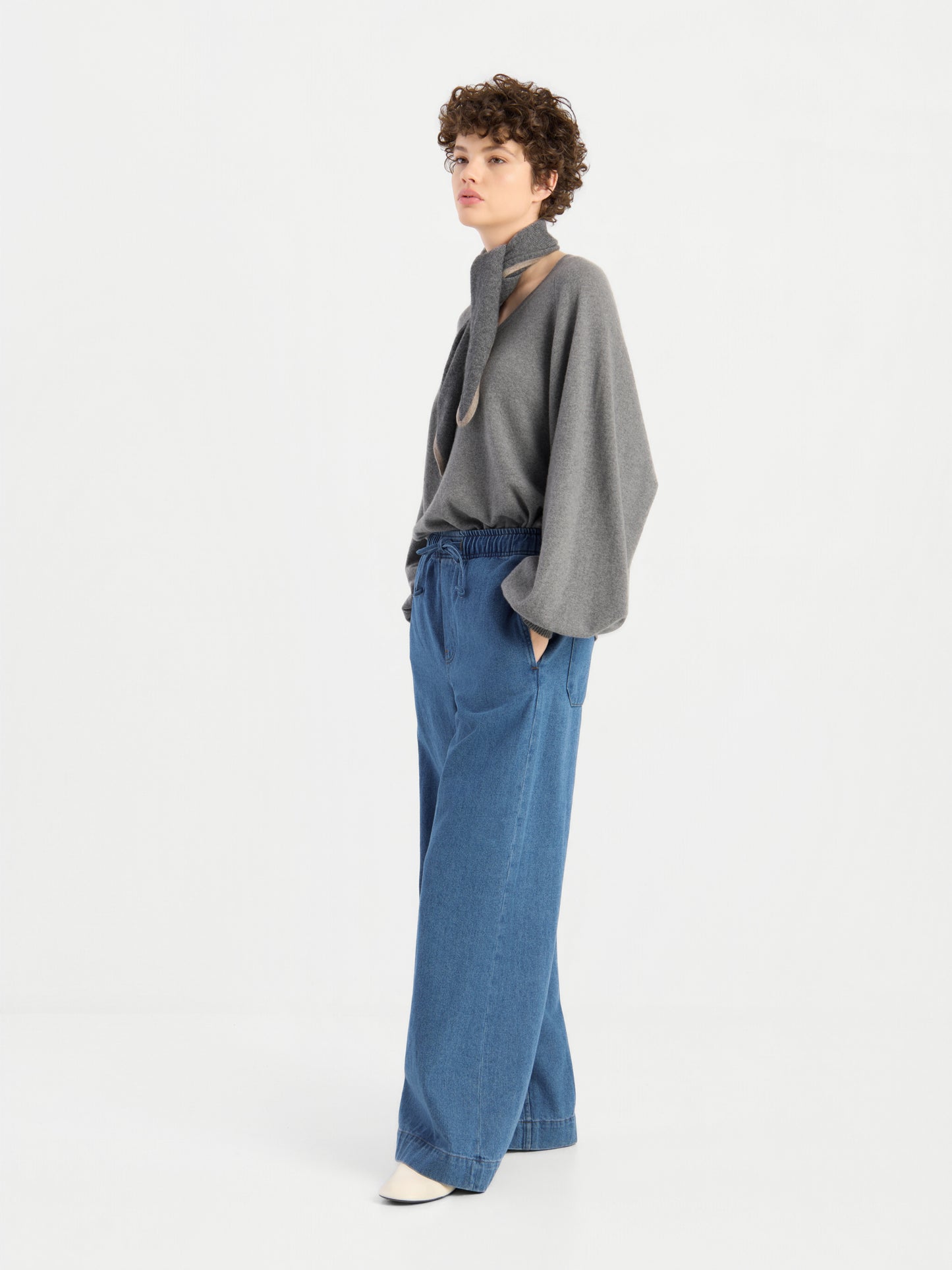Eine Person mit kurzen Locken trägt einen grauen Pullover mit weiten Ärmeln, eine „Anna Pants Denim“-Jeans, weiße Schuhe und einen braunen Schal und steht vor einem schlichten weißen Hintergrund.
