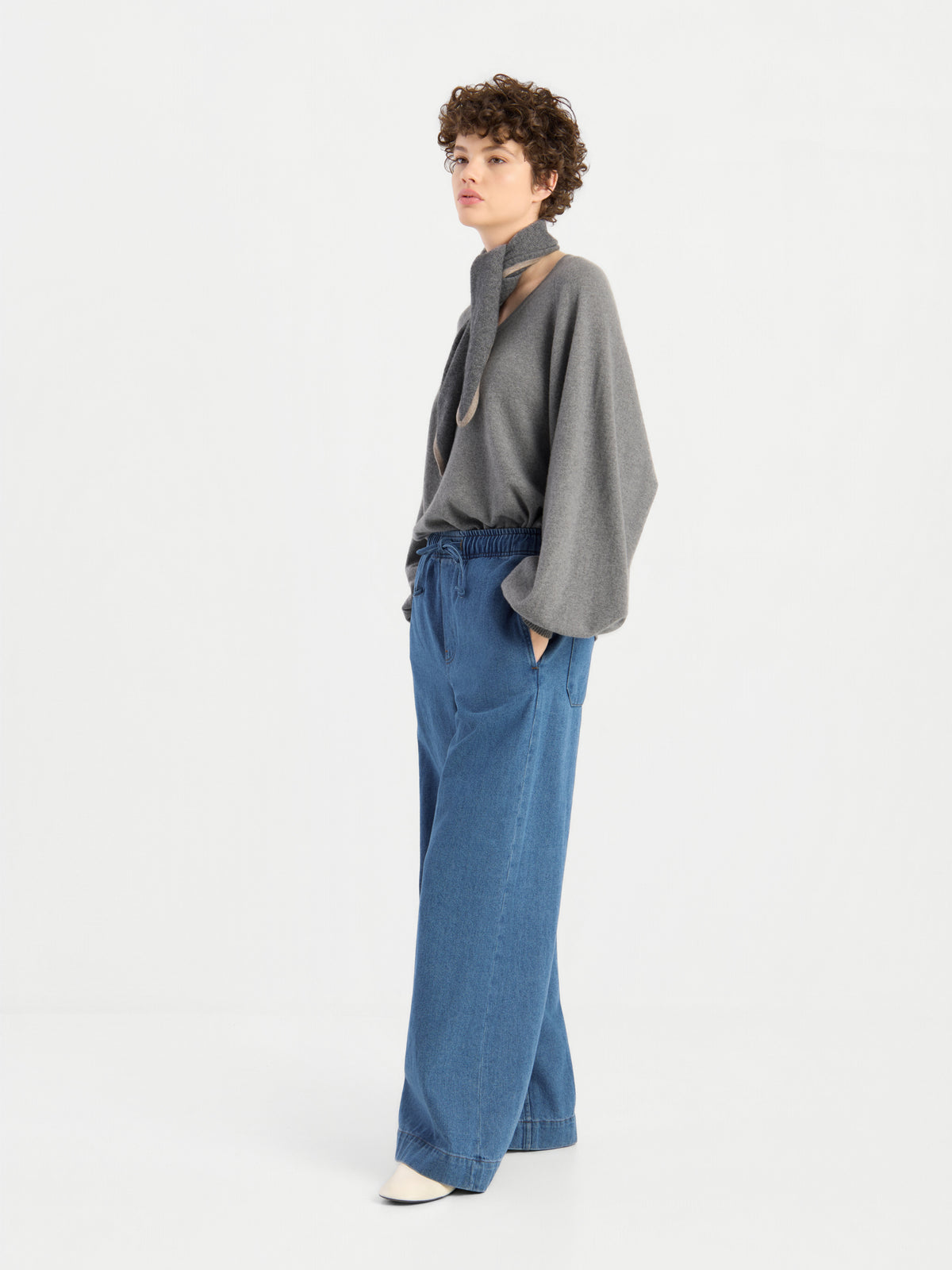 Ein Model, das den Pullover „Antonia V-Neck Grau“ mit weiten Ärmeln und eine blaue Jeans mit weitem Bein und Kordelzug trägt, kombiniert mit weißen Schuhen, steht vor einem hellgrauen Hintergrund.