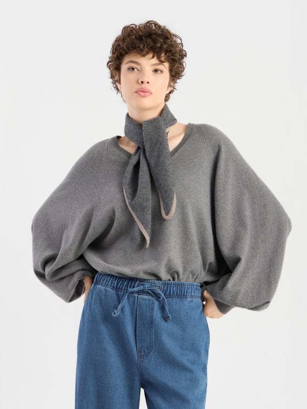 Eine Person trägt „Soft Volume“: einen grauen, übergroßen Pullover mit weiten Ärmeln, einen dazu passenden Schal, der am Hals gebunden ist, sowie eine blaue Hose mit Kordelzug; sie steht mit den Händen in den Taschen vor einem schlichten, hellen Hintergrund.
