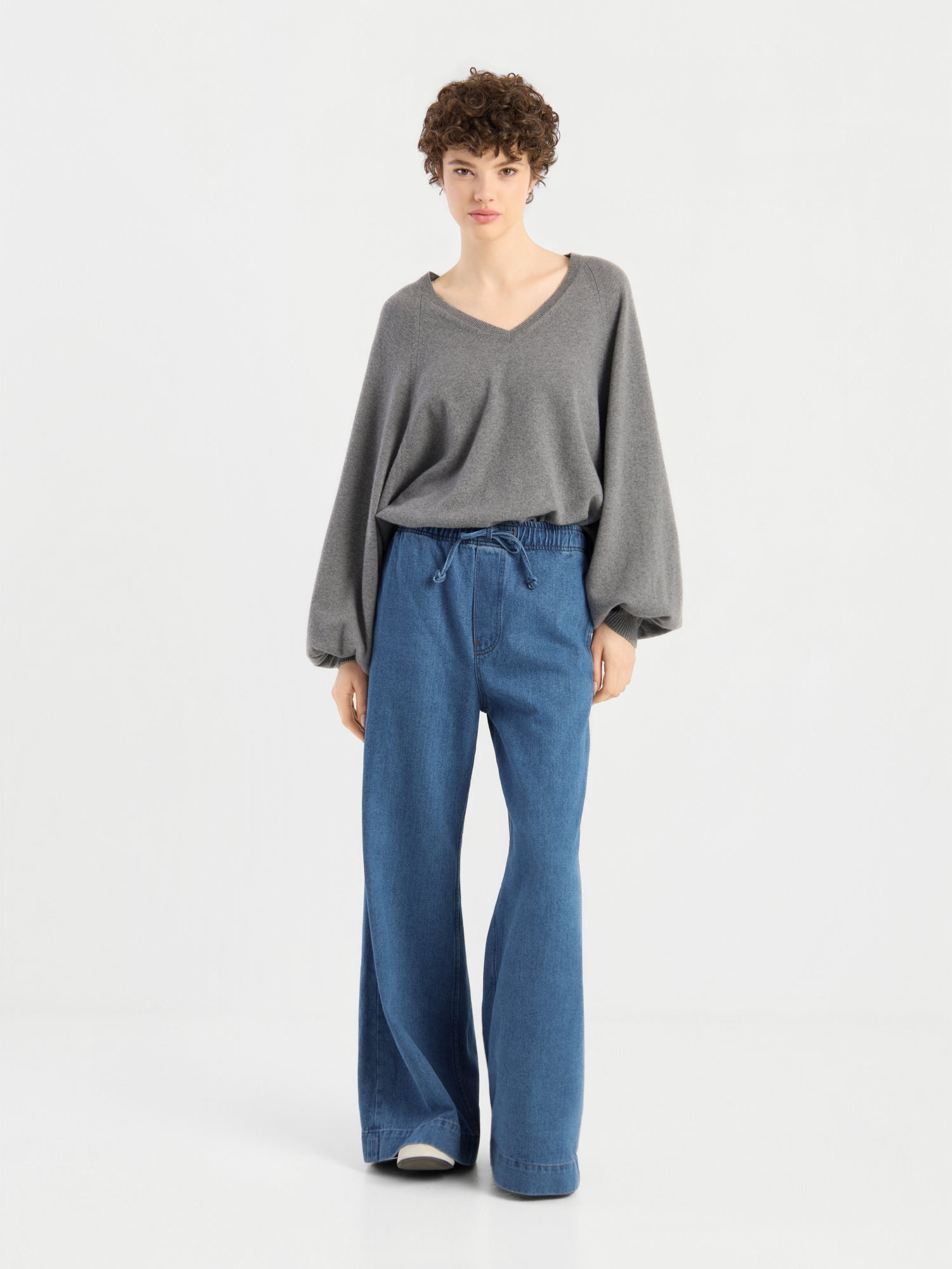 Eine Person mit kurzen, lockigen Haaren, die den Pullover „Antonia V-Neck Grau“ und eine blaue Jeans mit weitem Bein trägt und vor einem schlichten weißen Hintergrund steht.