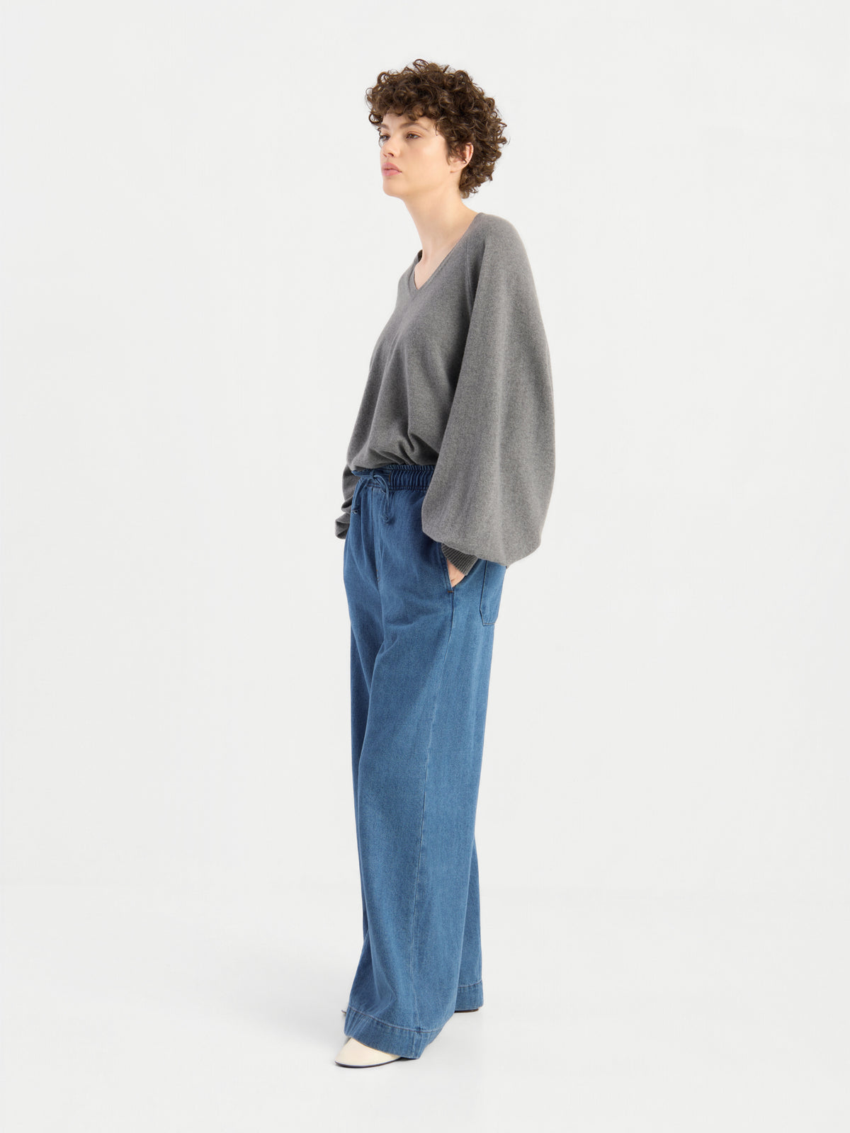 Ein weibliches Model mit kurzen Locken trägt den Pullover „Antonia“ mit V-Ausschnitt in Grau aus Merinowolle und eine blaue Jeans mit weitem Bein und Kordelzug und posiert vor einem weißen Hintergrund.
