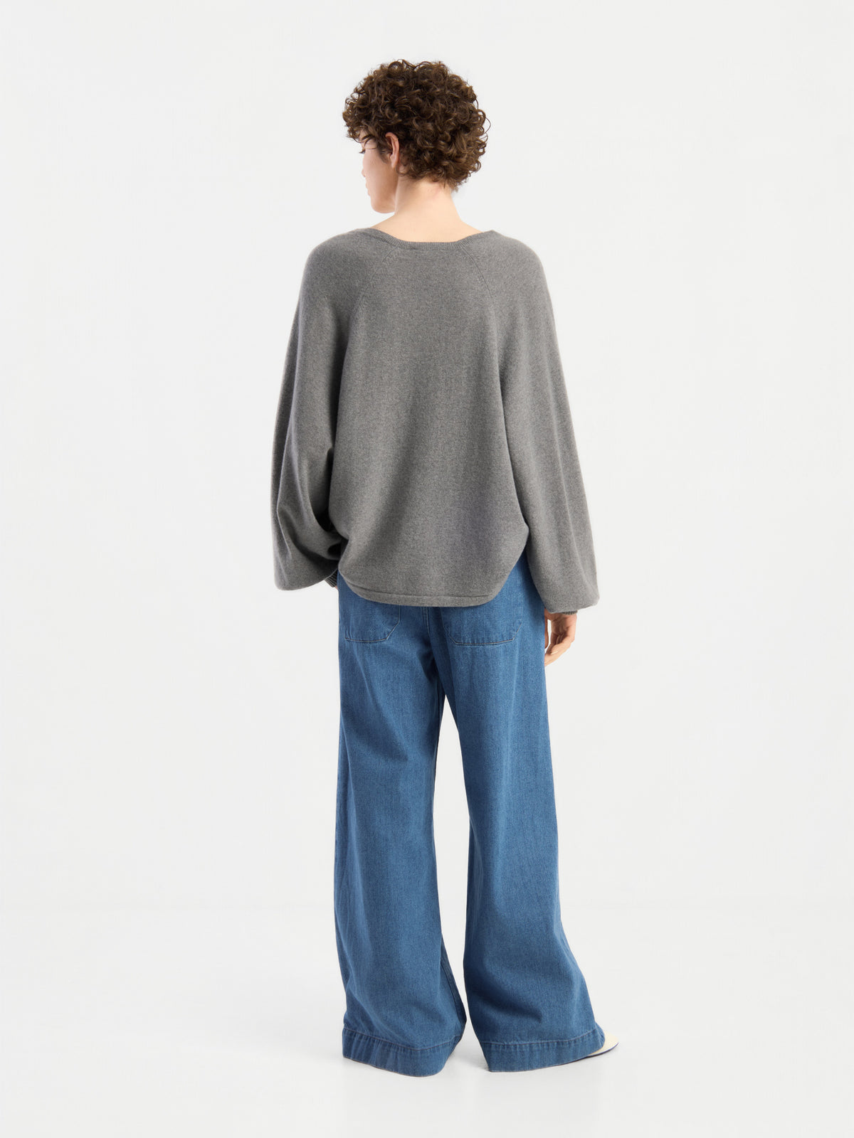 Eine Person mit kurzen, lockigen Haaren, von hinten gesehen, die einen locker sitzenden Pullover „Antonia V-Neck Grau“ und eine blaue Jeans mit weitem Bein trägt und vor einem schlichten weißen Hintergrund steht.