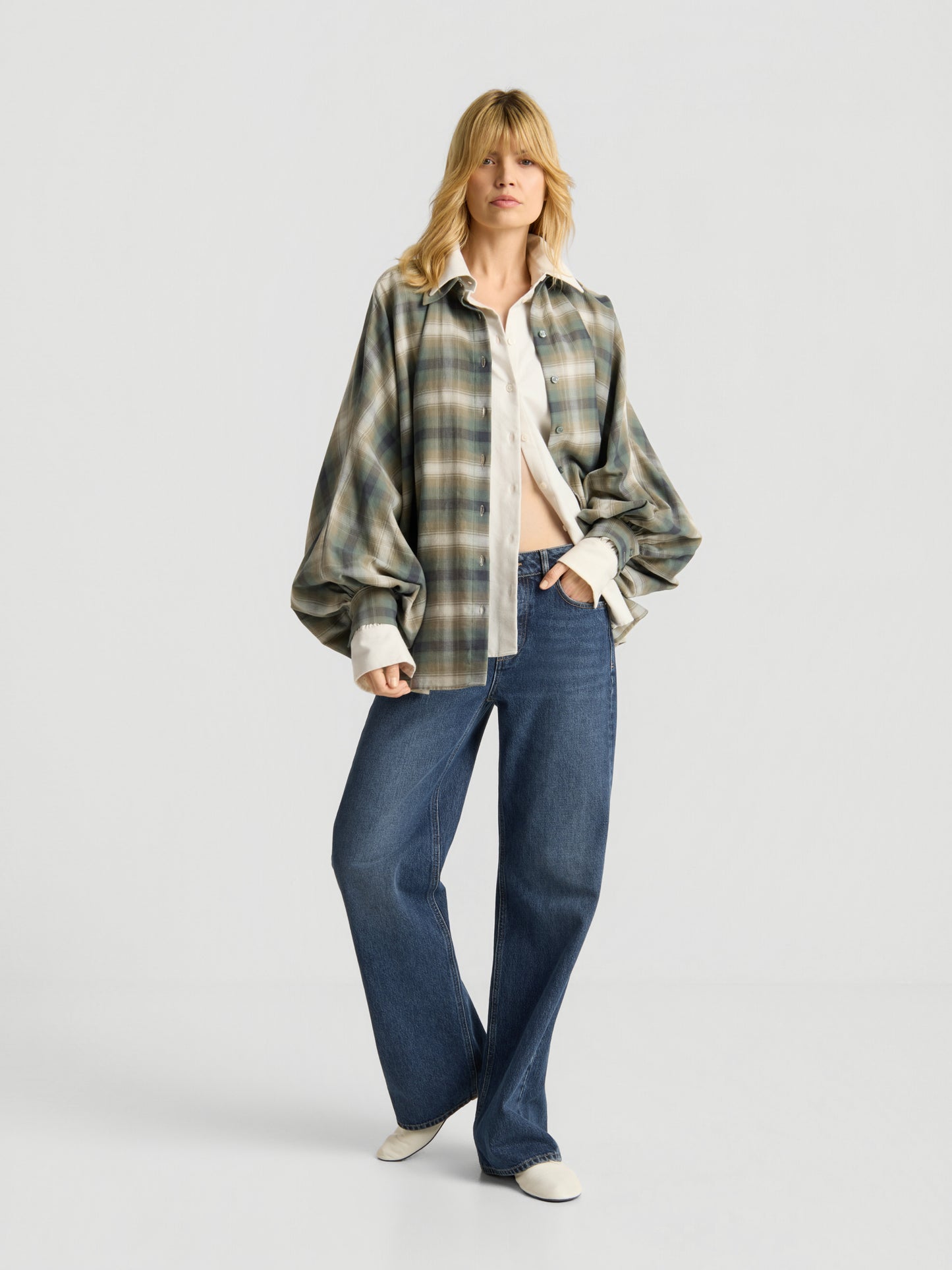 Eine Person mit blonden Haaren trägt die Bluse „Antonia Check Grün“ im Oversize-Look – ein kariertes Overshirt aus 100 % Baumwolle, das sowohl Komfort als auch Stil vereint –, über einem weißen Hemd, kombiniert mit einer blau-weit geschnittenen Jeans und weißen Schuhen, vor einem schlichten, hellen Hintergrund.