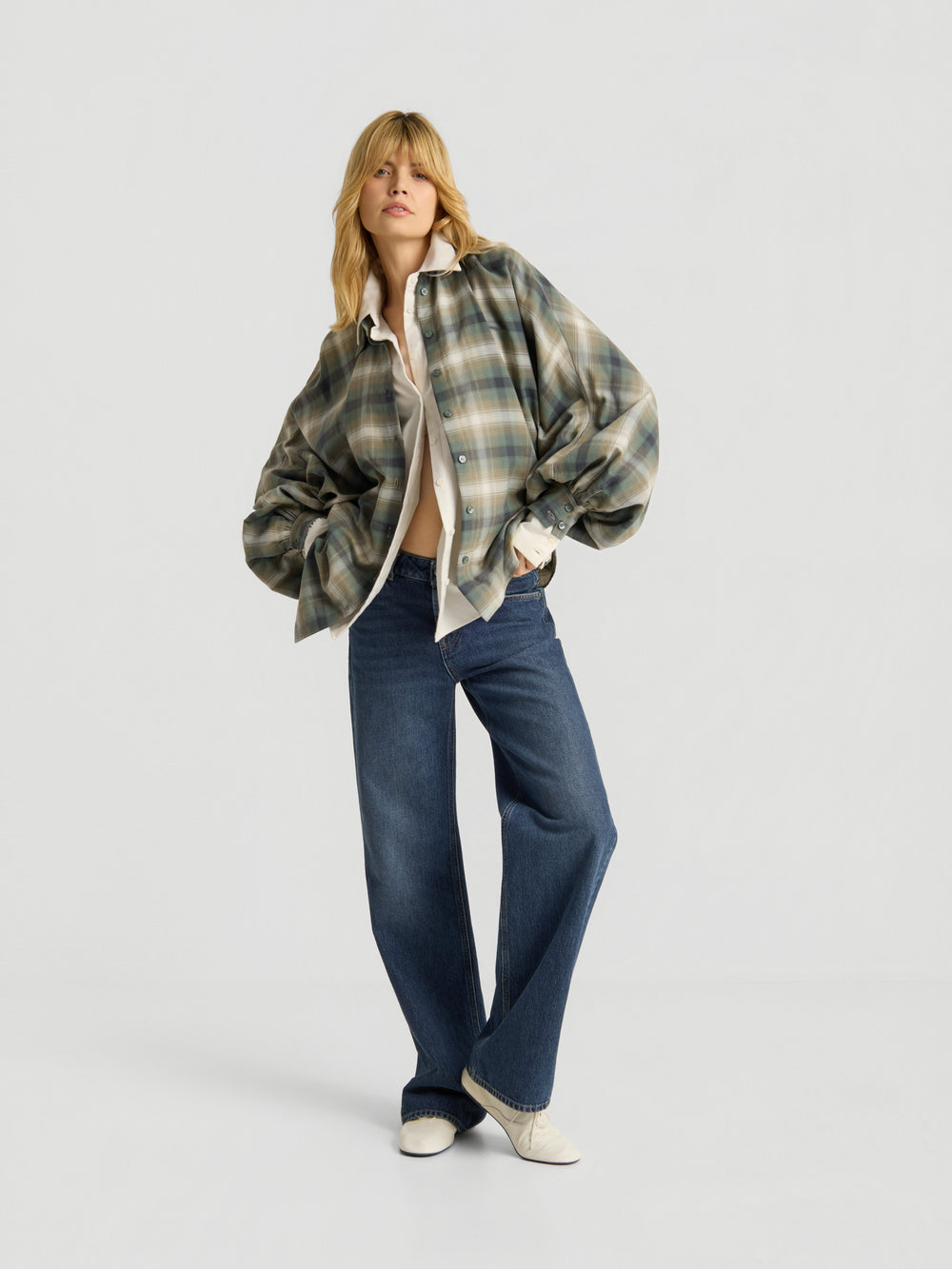 Eine Person mit langen blonden Haaren trägt das grüne karierte Oversize-Hemd von „Everyday Favorites“ und kombiniert es mit einer blauen Jeans mit weitem Bein und weißen Schuhen, während sie vor einem schlichten weißen Hintergrund steht.