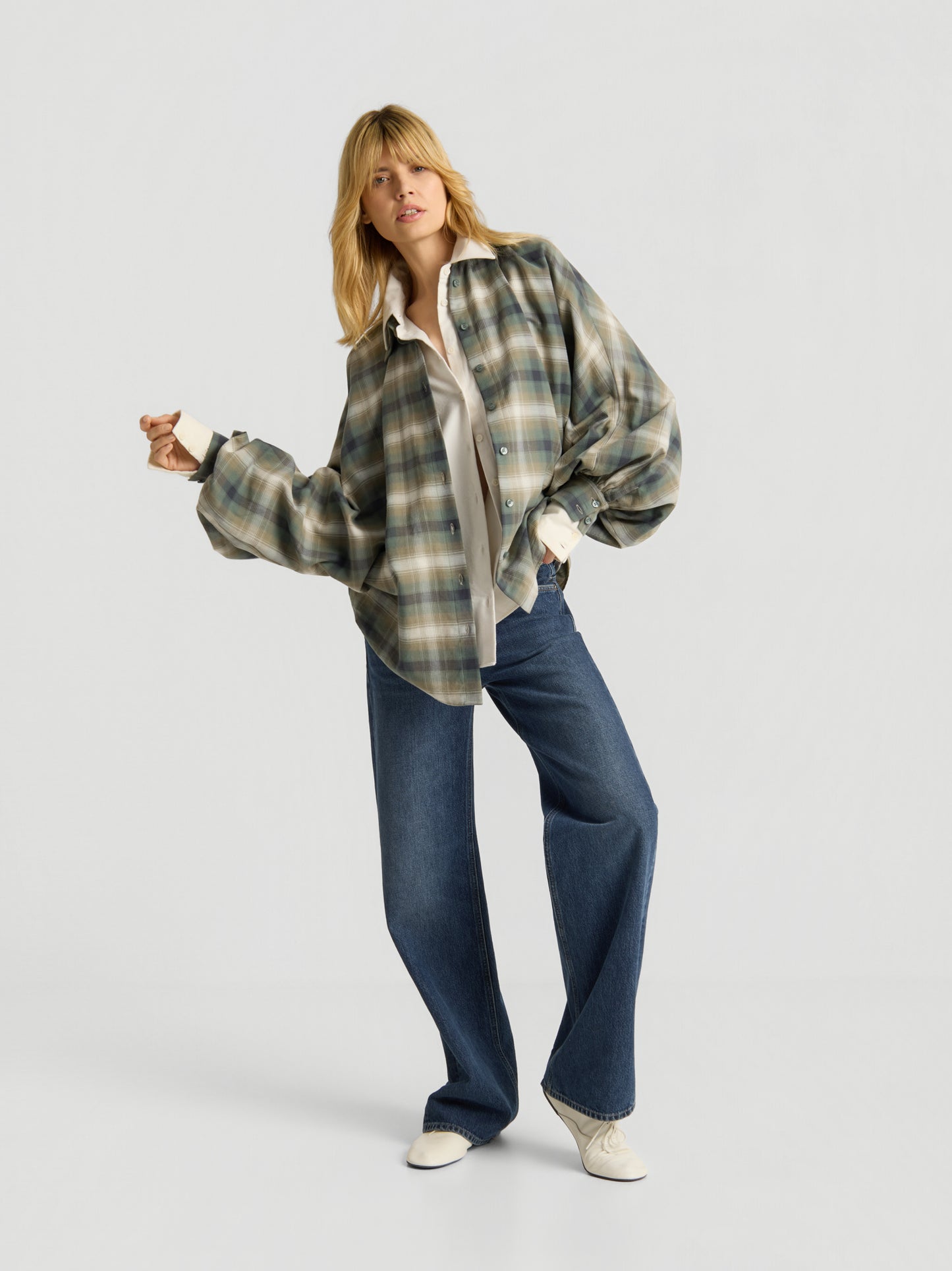 Eine Person mit blonden Haaren trägt die Bluse „Antonia Check Grün“ – eine karierte Damenbluse aus 100 % Baumwolle im Oversized-Look –, kombiniert mit einem weißen Shirt, blauen Wide-Leg-Jeans und weißen Schuhen, und posiert selbstbewusst vor einem schlichten, hellen Hintergrund.