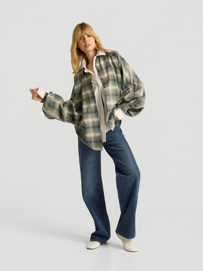 Eine Person mit blonden Haaren trägt die Bluse „Antonia Check Grün“ – eine karierte Damenbluse aus 100 % Baumwolle im Oversized-Look –, kombiniert mit einem weißen Shirt, blauen Wide-Leg-Jeans und weißen Schuhen, und posiert selbstbewusst vor einem schlichten, hellen Hintergrund.