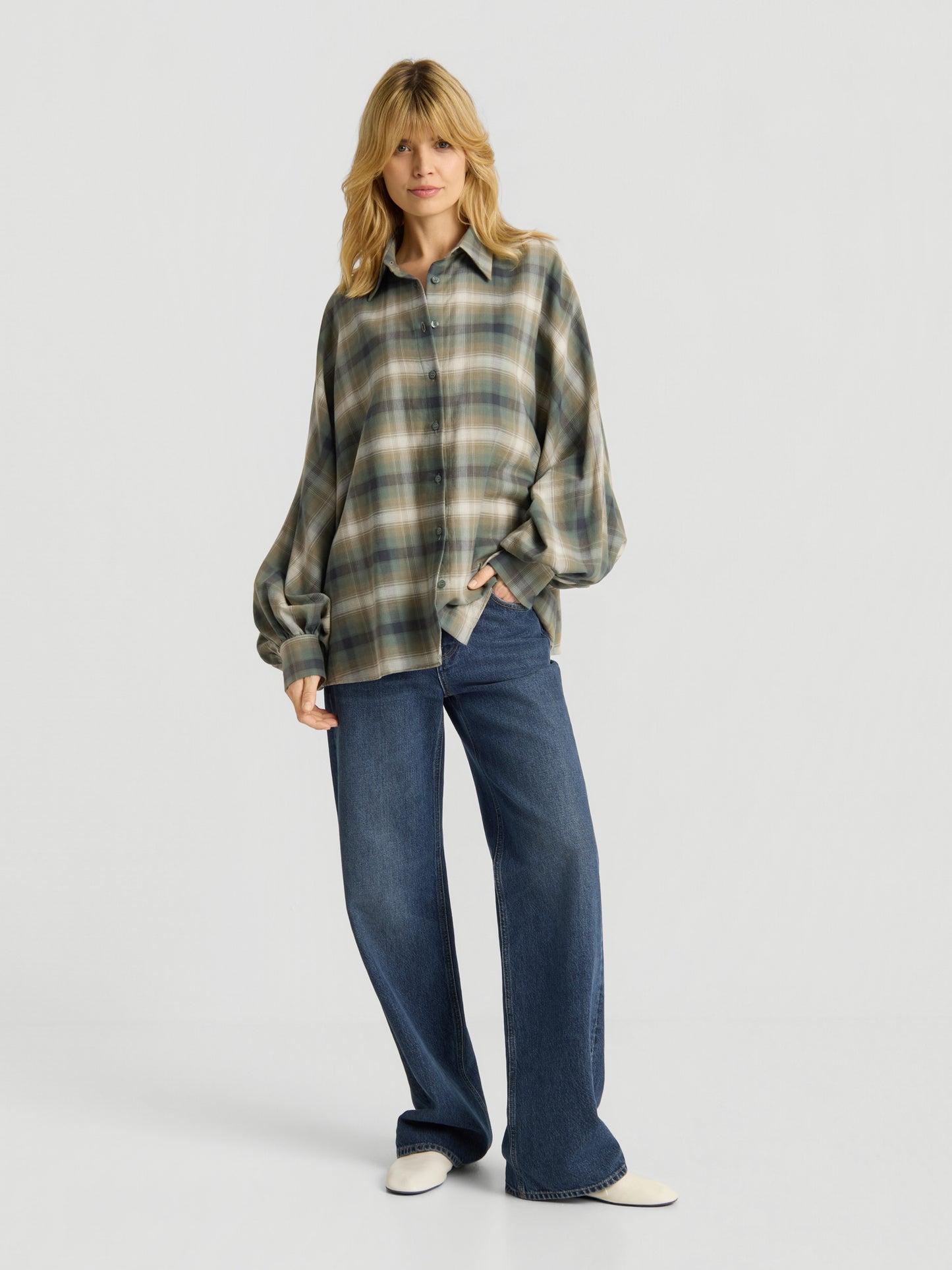 Eine Person mit langen blonden Haaren trägt die Bluse „Antonia Check Grün“ im Oversize-Look mit Ballonärmeln aus 100 % Baumwolle, kombiniert mit einer blau-weit geschnittenen Jeans und weißen Schuhen, und steht vor einem schlichten weißen Hintergrund.