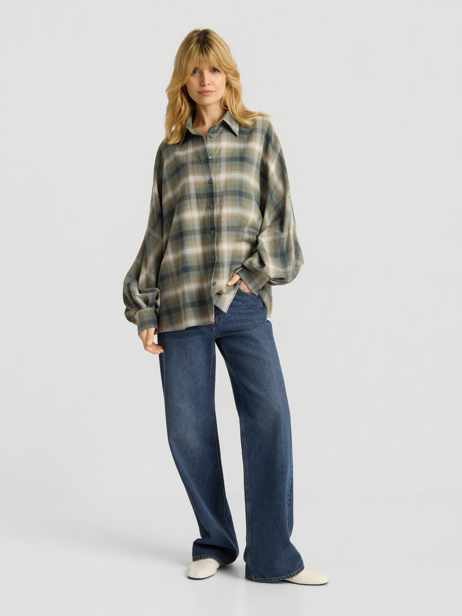 Eine Person mit langen blonden Haaren trägt die Bluse „Antonia Check Grün“ im Oversize-Look mit Ballonärmeln aus 100 % Baumwolle, kombiniert mit einer blau-weit geschnittenen Jeans und weißen Schuhen, und steht vor einem schlichten weißen Hintergrund.