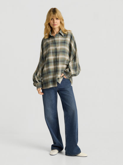 Eine Person mit langen blonden Haaren trägt die Bluse „Antonia Check Grün“ im Oversize-Look mit Ballonärmeln aus 100 % Baumwolle, kombiniert mit einer blau-weit geschnittenen Jeans und weißen Schuhen, und steht vor einem schlichten weißen Hintergrund.