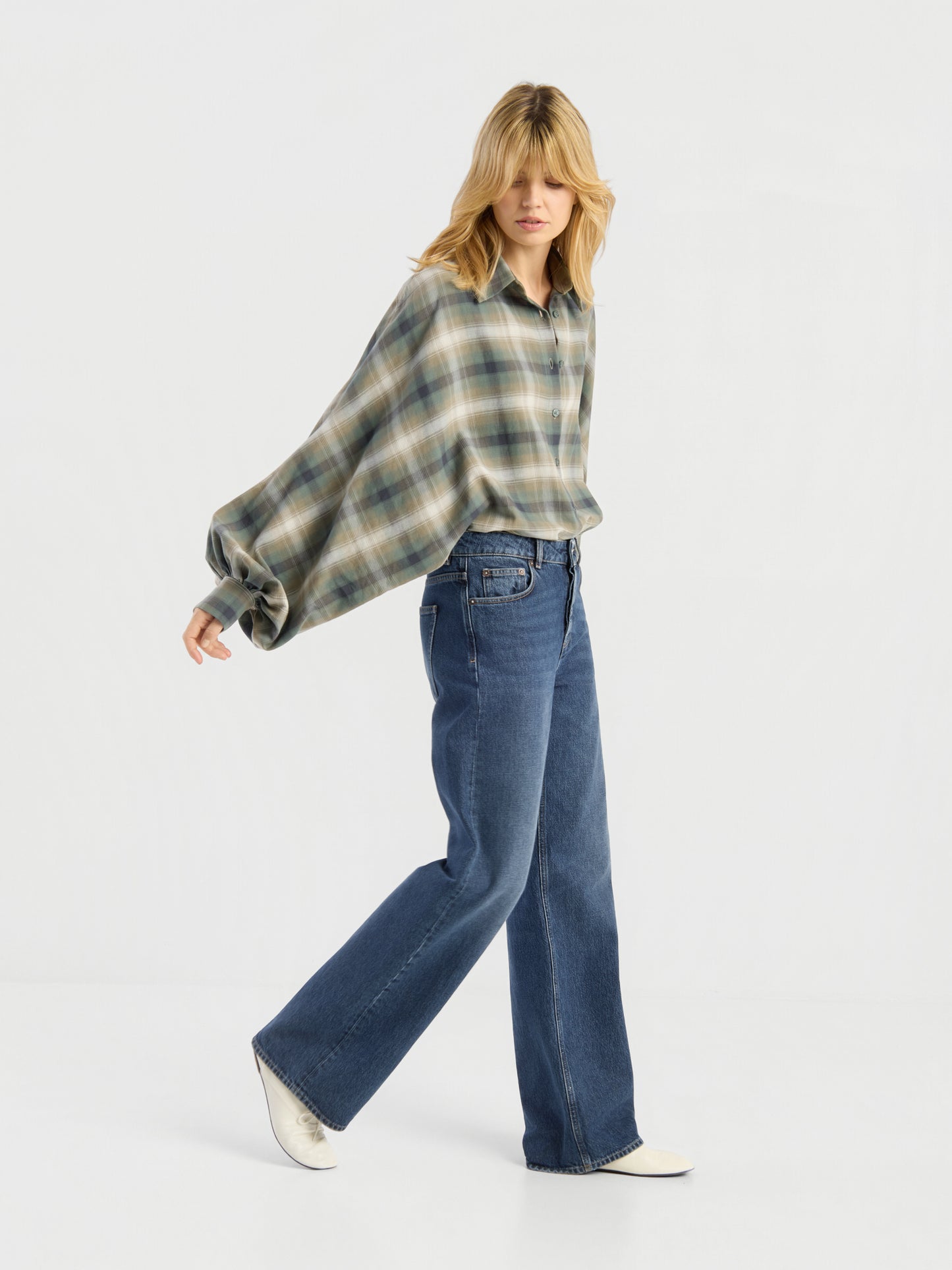 Eine Person mit blonden Haaren läuft in einem Studio vor einem schlichten weißen Hintergrund und trägt eine Bluse „Antonia Check Grün“ aus 100 % Baumwolle im Oversize-Look sowie eine blaue Jeans mit weitem Bein, dazu weiße Schuhe.