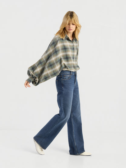 Eine Person mit blonden Haaren läuft in einem Studio vor einem schlichten weißen Hintergrund und trägt eine Bluse „Antonia Check Grün“ aus 100 % Baumwolle im Oversize-Look sowie eine blaue Jeans mit weitem Bein, dazu weiße Schuhe.