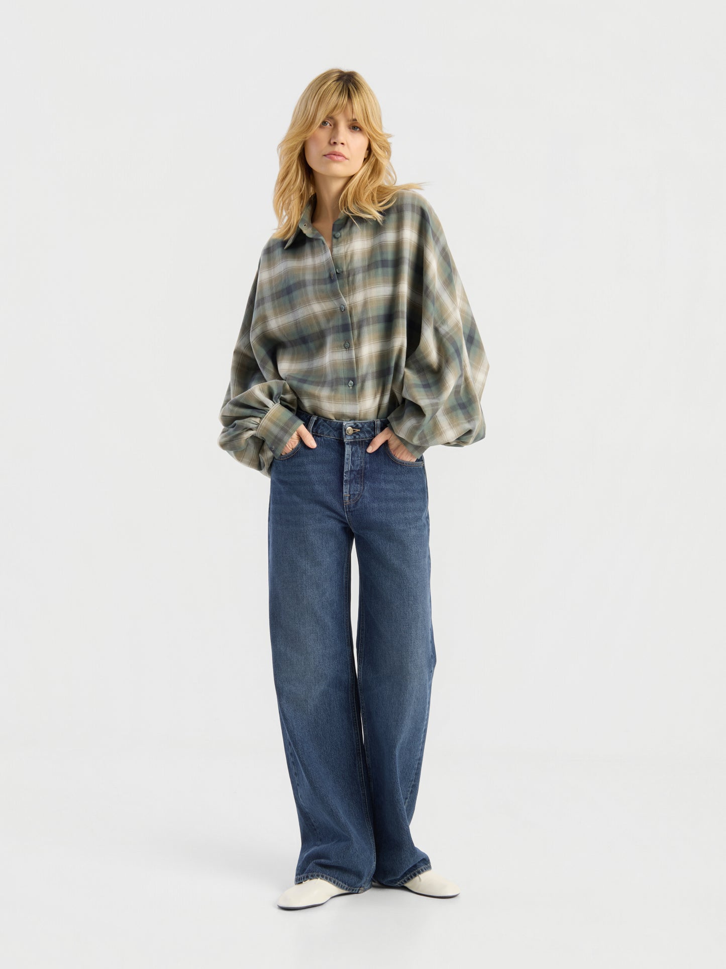 Eine Frau mit blonden Haaren trägt die Bluse „Antonia Check Grün“ im Oversize-Look – eine grün-grau karierte Damenbluse mit Ballonärmeln aus 100 % Baumwolle –, dazu eine blaue Jeans mit weitem Bein und weiße Schuhe; sie steht vor einem schlichten weißen Hintergrund.