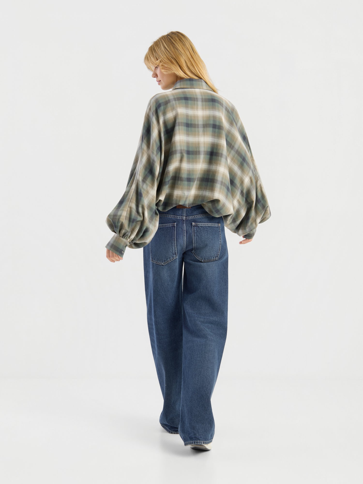 Eine Person mit langen blonden Haaren, die eine Bluse im Oversize-Look „Antonia Check Grün“ trägt – eine grüne, karierte Damenbluse aus 100 % Baumwolle mit weiten Ärmeln – sowie eine lockere blaue Jeans, geht vor einem schlichten weißen Hintergrund von der Kamera weg.