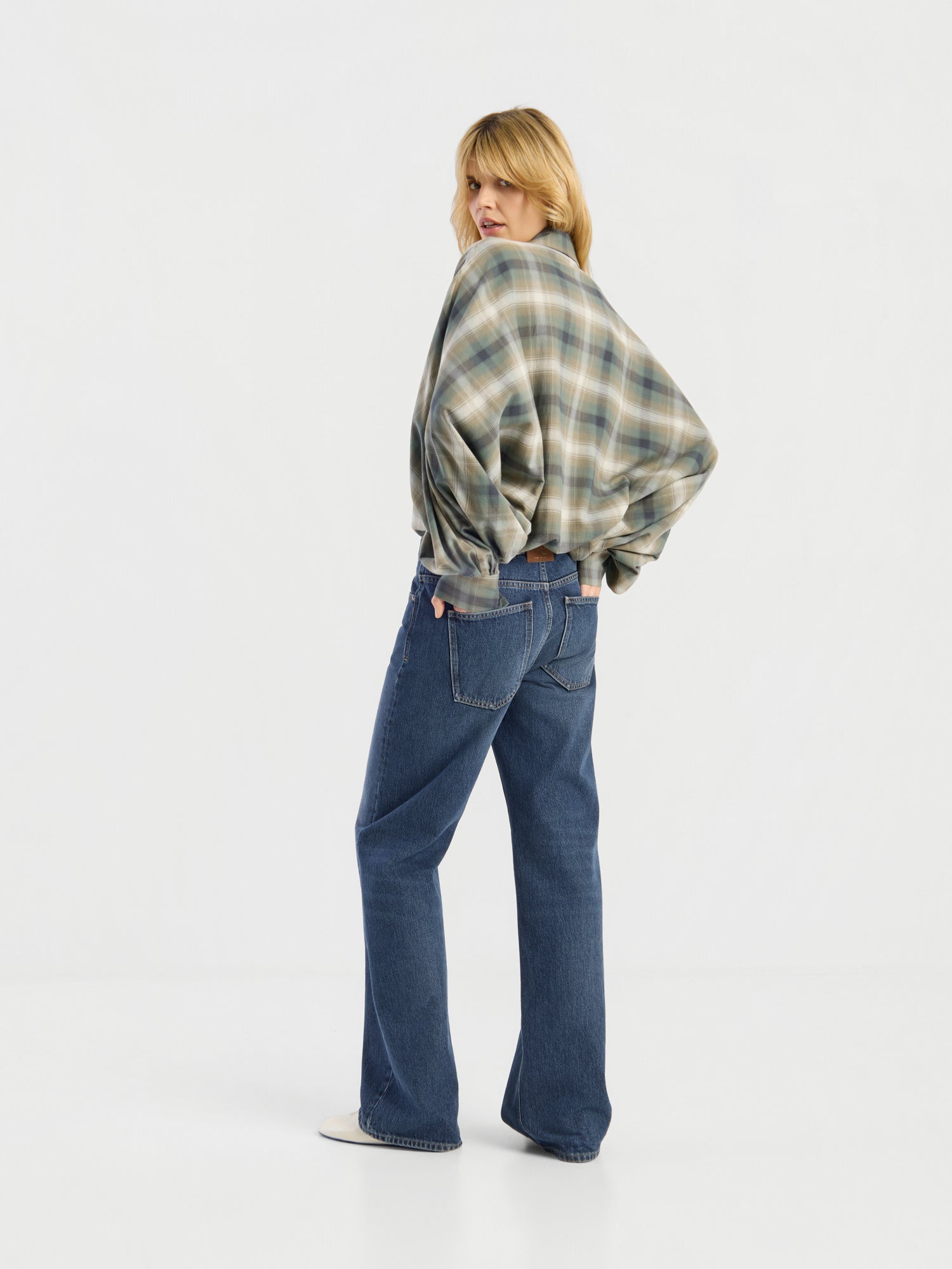 Eine Person, die die Bluse „Antonia Check Grün“ und eine blaue Jeans trägt, steht in einem weißen Studio, mit dem Rücken zur Kamera, dreht jedoch den Kopf, um über die Schulter zurückzuschauen. Der Stoff der Bluse aus 100 % Baumwolle fällt angenehm und verleiht der Szene einen lässigen Stil.