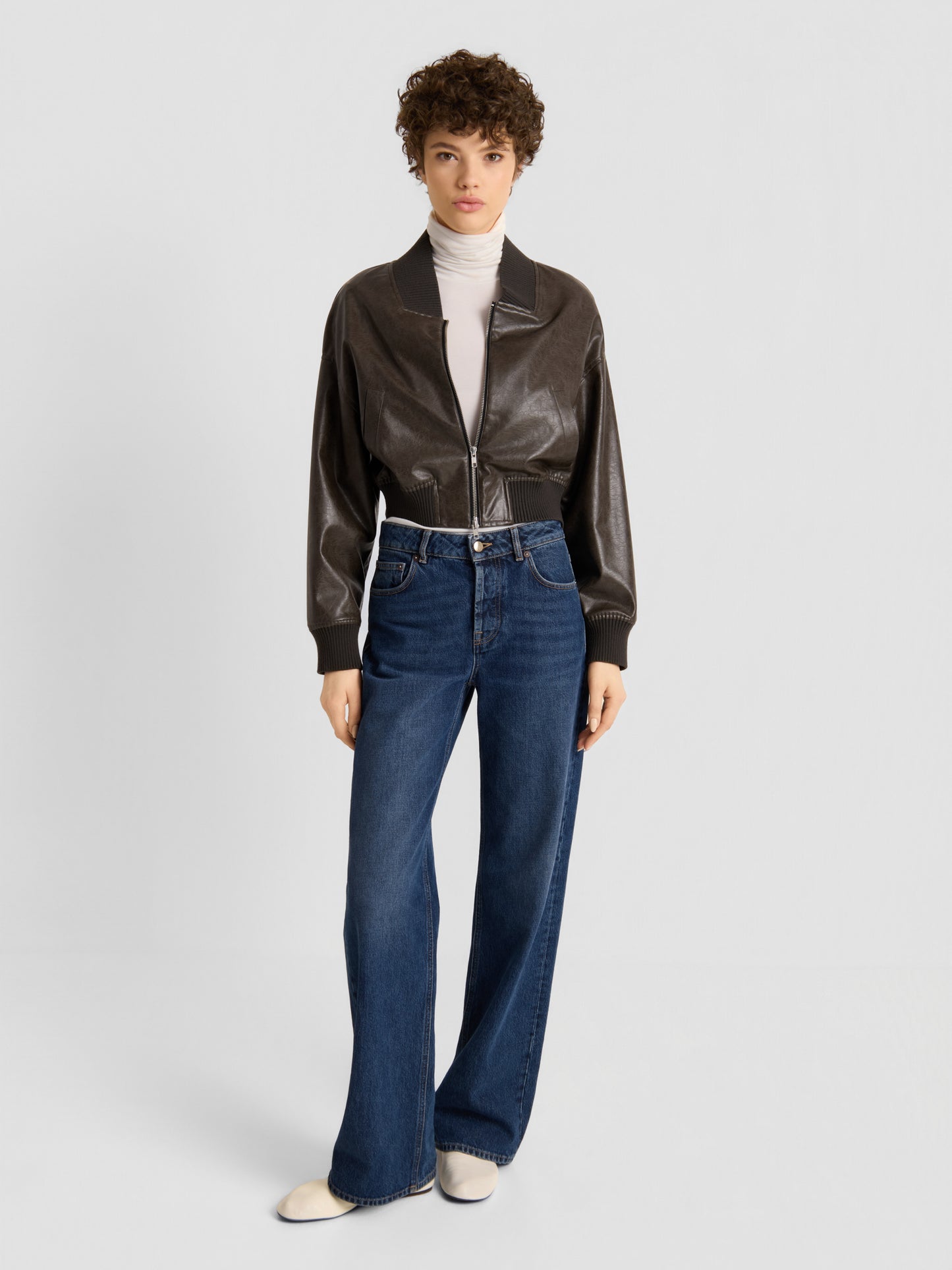 Eine Person, die die „Bomber Vegan Leather“-Jacke, einen weißen Rollkragenpullover, eine blaue Jeans mit weitem Bein und weiße Schuhe trägt, steht vor einem schlichten, hellen Hintergrund und blickt mit neutralem Gesichtsausdruck nach vorne.