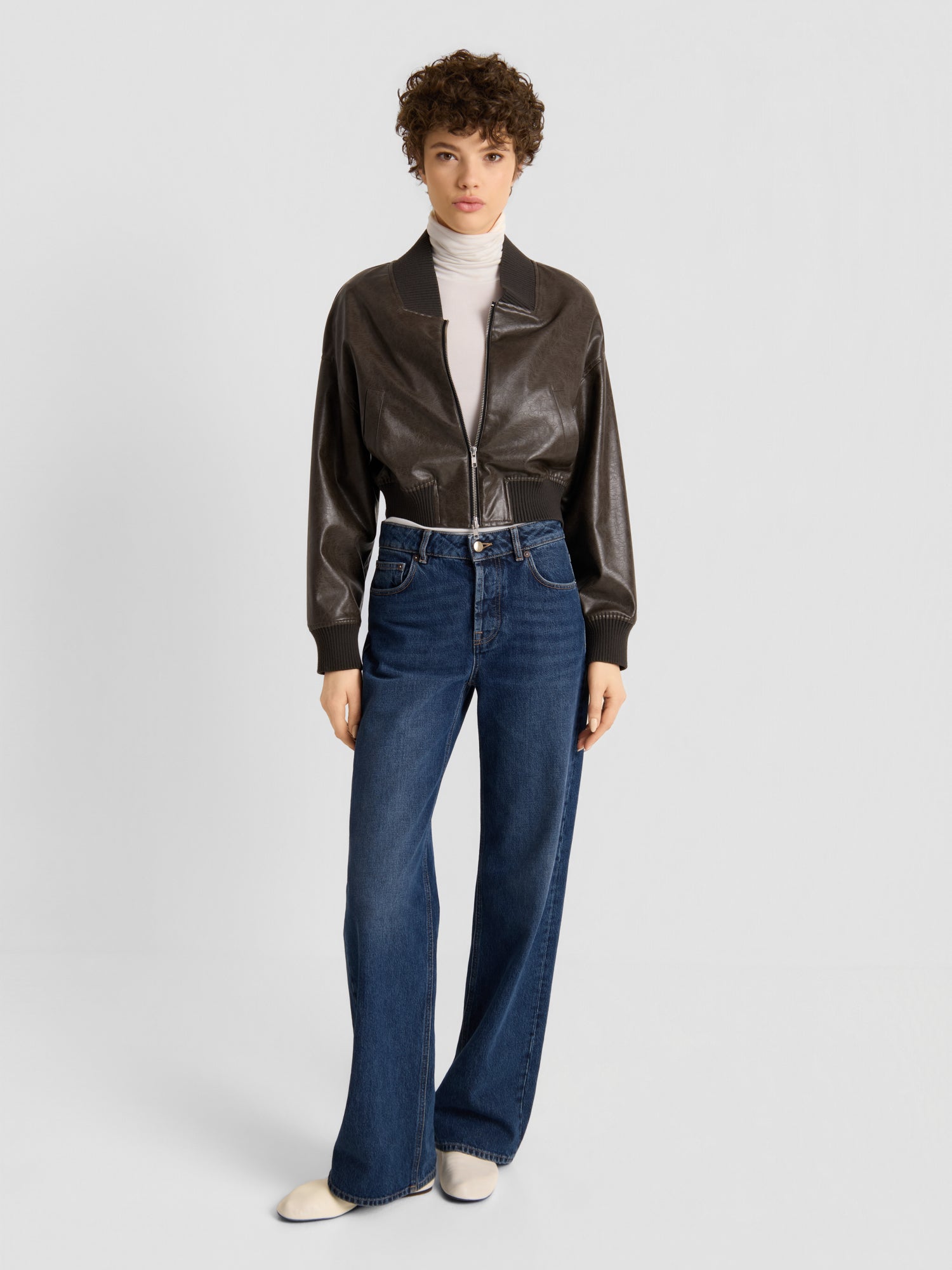 Eine Person, die die „Bomber Vegan Leather“-Jacke, einen weißen Rollkragenpullover, eine blaue Jeans mit weitem Bein und weiße Schuhe trägt, steht vor einem schlichten, hellen Hintergrund und blickt mit neutralem Gesichtsausdruck nach vorne.