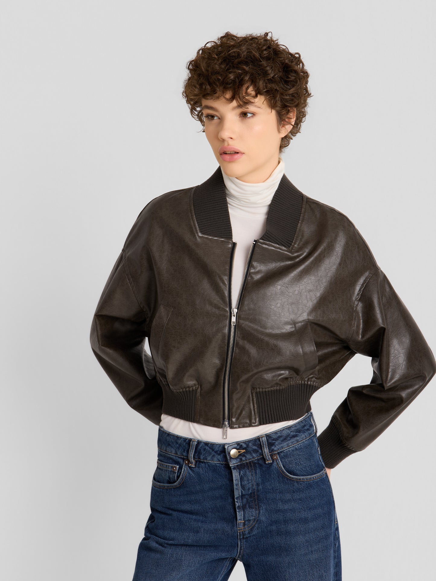 Ein Model präsentiert die „Bomber“-Jacke aus veganem Leder in Dunkelbraun, die über einem weißen Rollkragenpullover und zu blauen Jeans getragen wird, und steht mit den Händen in den Taschen vor einem schlichten, hellen Hintergrund.