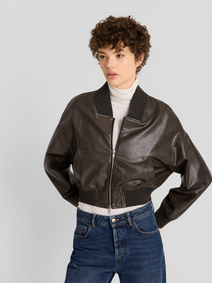 Ein Model präsentiert die „Bomber“-Jacke aus veganem Leder in Dunkelbraun, die über einem weißen Rollkragenpullover und zu blauen Jeans getragen wird, und steht mit den Händen in den Taschen vor einem schlichten, hellen Hintergrund.