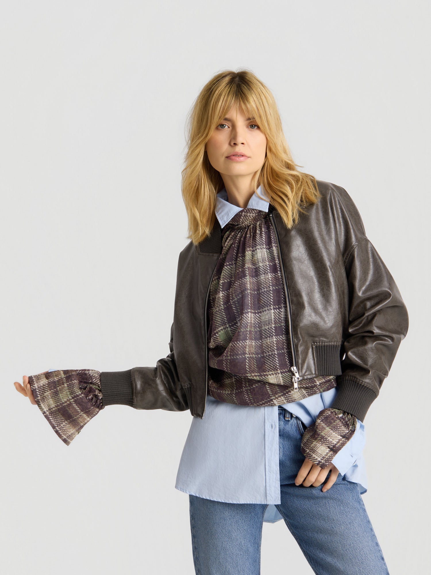 Eine Frau mit blonden Haaren posiert vor einem schlichten hellgrauen Hintergrund. Sie trägt die „Bomber Vegan Leather“-Jacke in Braun, darüber ein blaues Hemd und einen karierten Schal, dazu eine Jeans. Sie steht mit angewinkeltem Arm da.