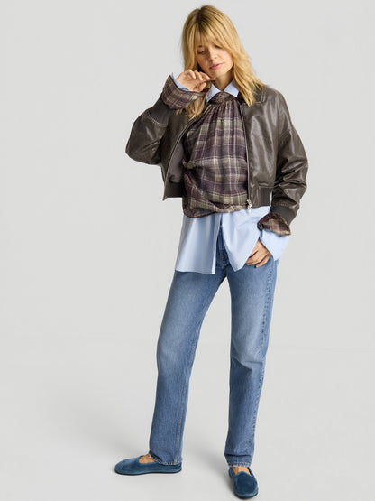 Eine Frau steht in Freizeitkleidung da: blaue Jeans, blaue Slipper, ein blaues Hemd, darüber ein kariertes Oberteil, und die „Bomber“-Jacke aus veganem Leder. Sie hat blondes Haar, ihre Augen sind geschlossen, und eine Hand hält sie in der Nähe ihres Gesichts.