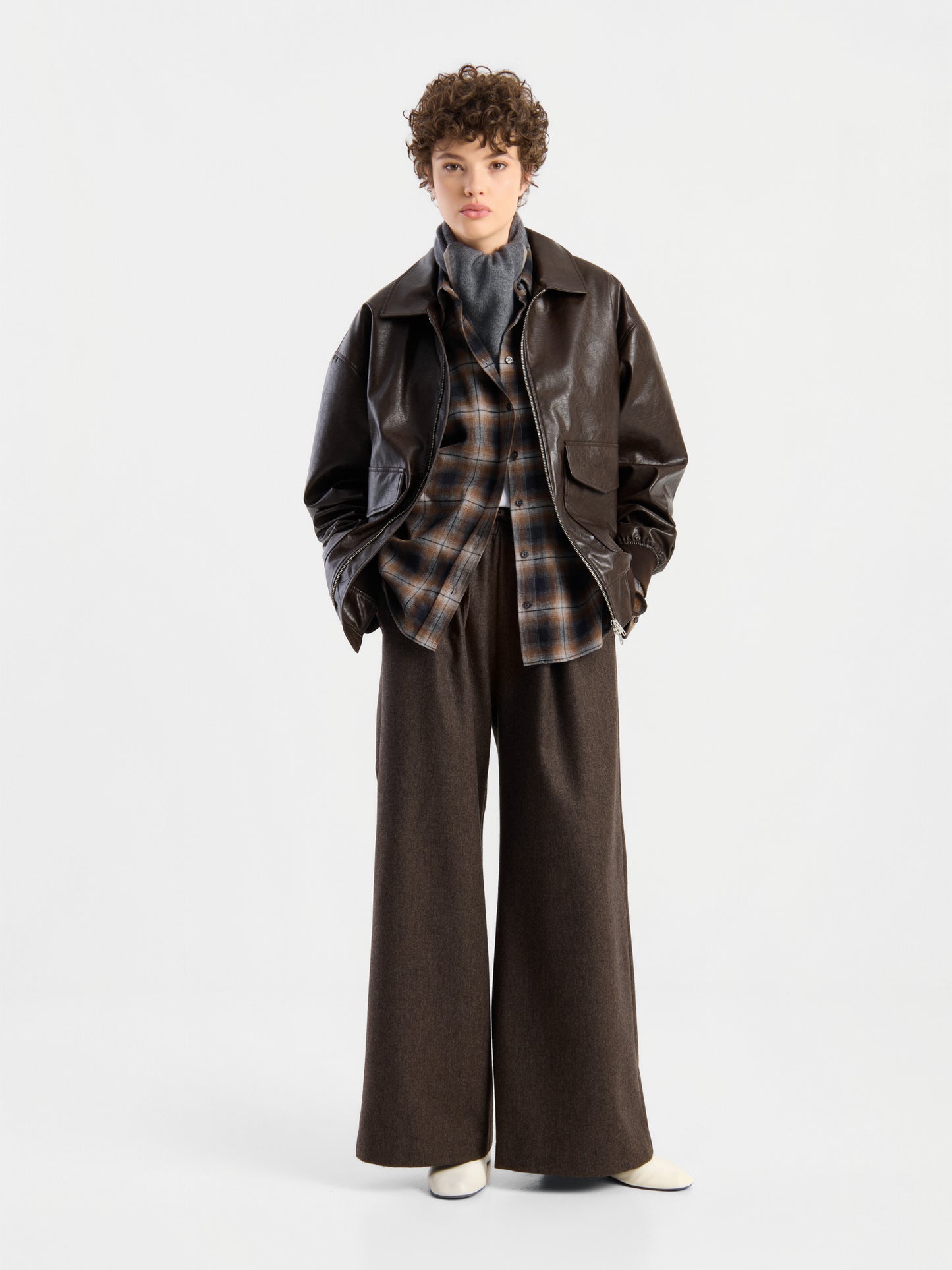 Eine Person mit lockigem Haar trägt eine braune Oversize-Jacke aus veganem Leder über einem karierten Hemd, einem grauen Rollkragenpullover, einer weiten braunen Hose und weißen Schuhen und steht vor einem schlichten weißen Hintergrund.