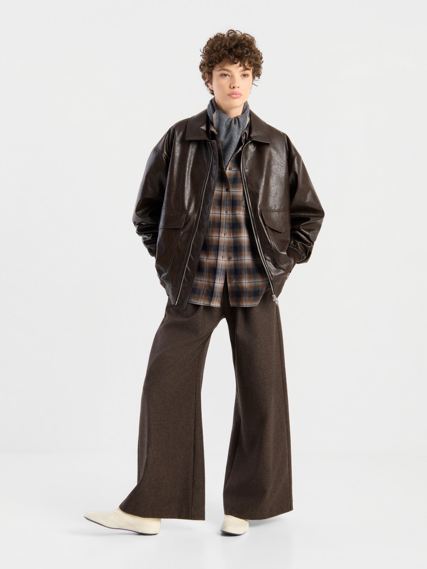 Eine Person steht vor einem weißen Hintergrund und trägt die Oversized-Jacke aus veganem Leder in Braun, kombiniert mit einem karierten Hemd, einem grauen Rollkragenpullover, einer braunen Hose mit weitem Bein und weißen Schuhen. Die Hände stecken in den Taschen, der Gesichtsausdruck ist neutral.