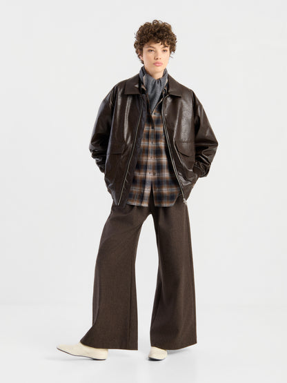 Eine Person steht vor einem weißen Hintergrund und trägt die Oversized-Jacke aus veganem Leder in Braun, kombiniert mit einem karierten Hemd, einem grauen Rollkragenpullover, einer braunen Hose mit weitem Bein und weißen Schuhen. Die Hände stecken in den Taschen, der Gesichtsausdruck ist neutral.
