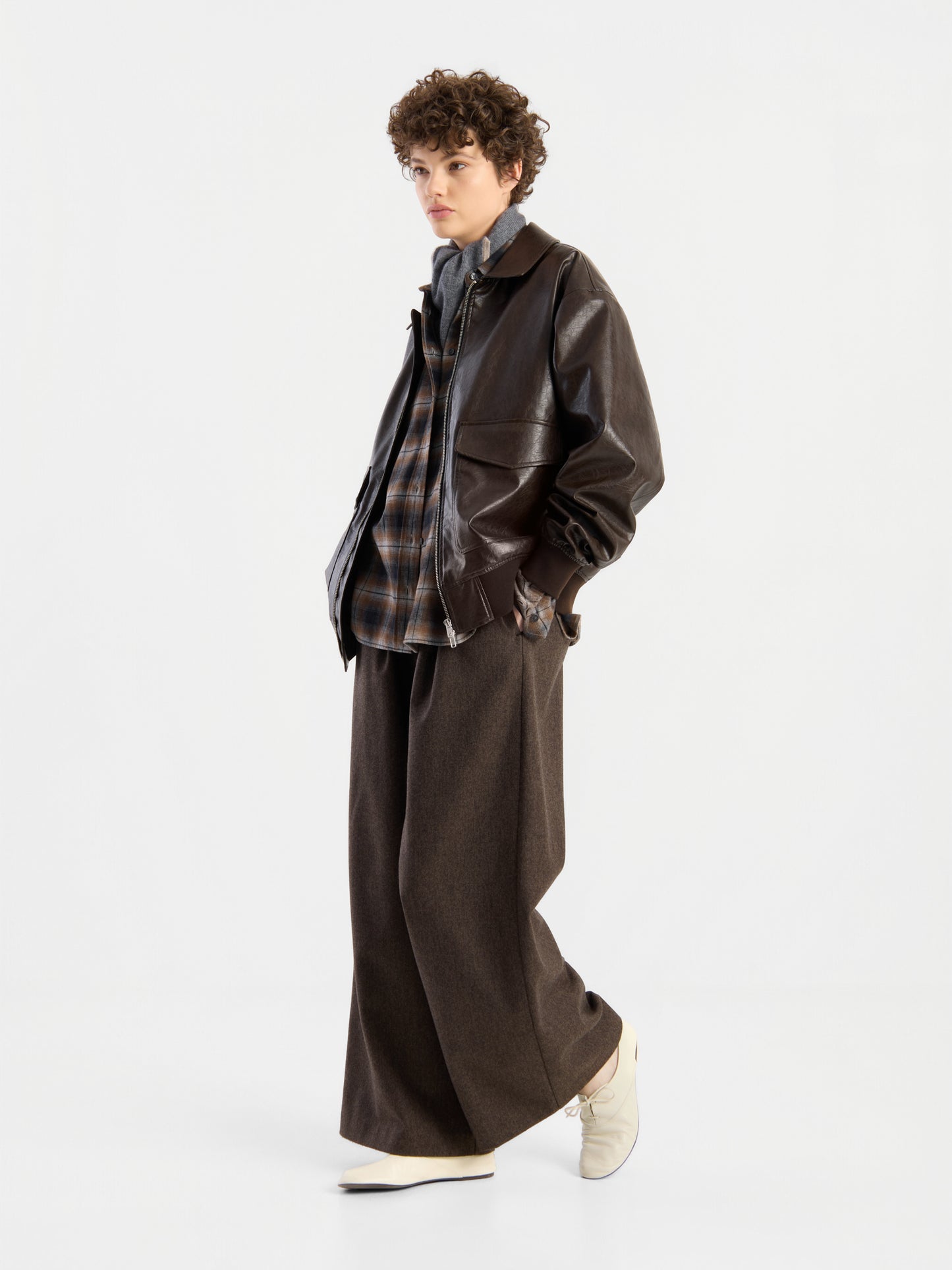 Eine Person mit lockigem Haar, die eine braune Oversize-Jacke aus veganem Leder über einem karierten Hemd, einer braunen Hose mit weitem Bein und weißen Schuhen trägt und vor einem schlichten weißen Hintergrund steht.