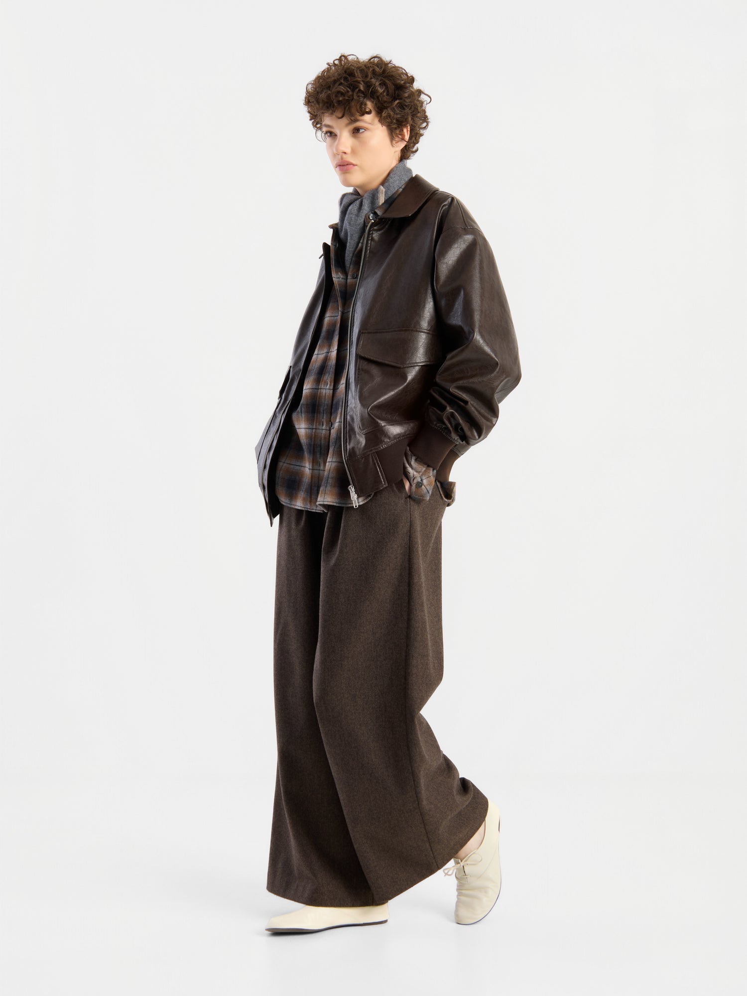 Eine Person mit lockigem Haar, die eine braune Oversize-Jacke aus veganem Leder über einem karierten Hemd, einer braunen Hose mit weitem Bein und weißen Schuhen trägt und vor einem schlichten weißen Hintergrund steht.