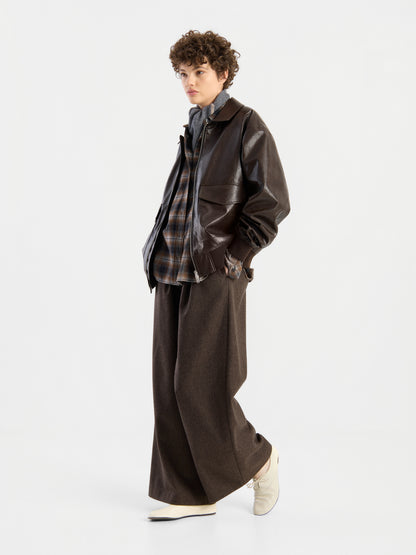 Eine Person mit lockigem Haar, die eine braune Oversize-Jacke aus veganem Leder über einem karierten Hemd, einer braunen Hose mit weitem Bein und weißen Schuhen trägt und vor einem schlichten weißen Hintergrund steht.