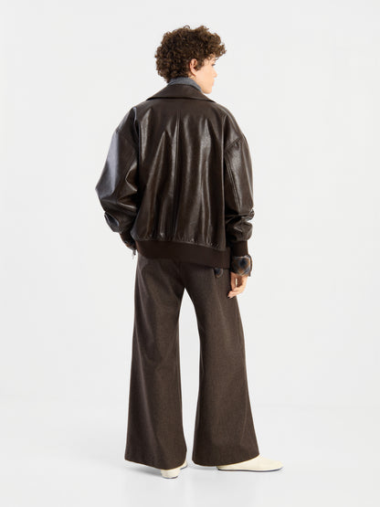 Eine Person mit kurzen Locken steht mit dem Rücken zur Kamera und trägt die Oversized-Jacke aus veganem Leder in Dunkelbraun, kombiniert mit einer braunen Hose mit weitem Bein und weißen Schuhen vor einem schlichten weißen Hintergrund.